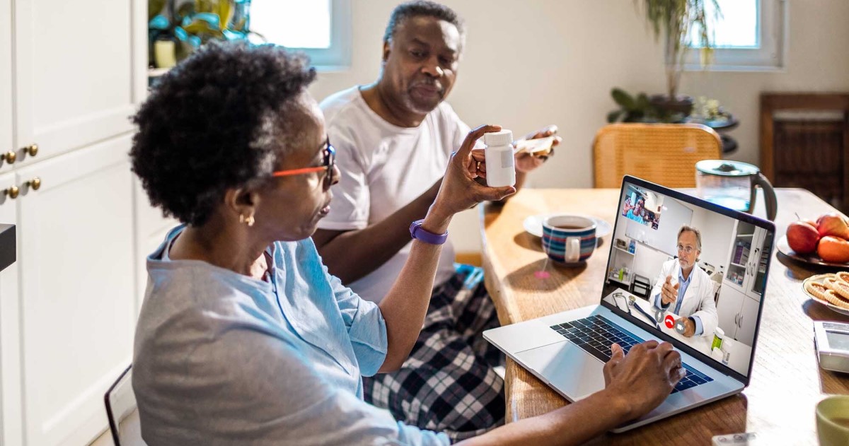 Person on laptop at kitchen table with another who holds up prescription bottle for telehealth doctor