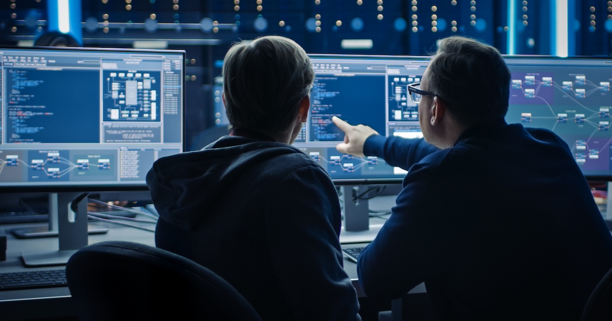 Two individuals sitting in front of two computer monitors with their backs turned toward the camera