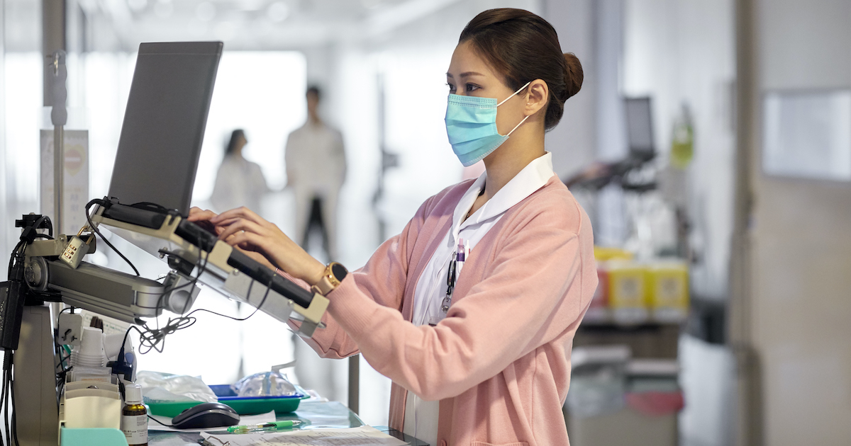 A nurse checking a patient's file on a laptop mounted in a hospital corridor