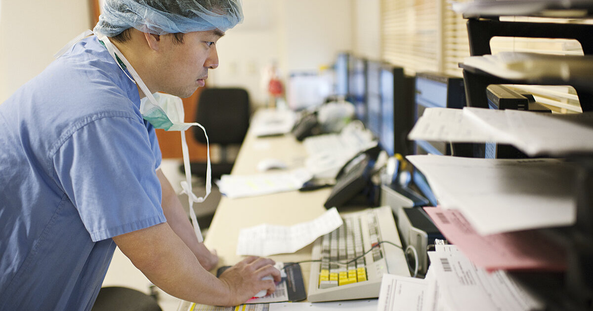 A clinician wearing scrubs is looking at a patient record from a computer screen