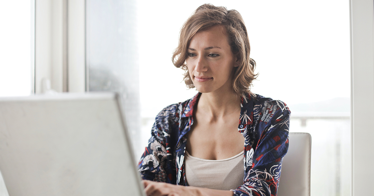 Woman sits at a table and is reading and typing on a laptop