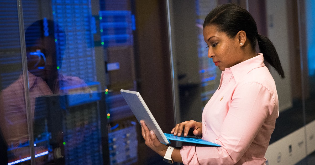 A person stands with a laptop in front of a stack of computer servers