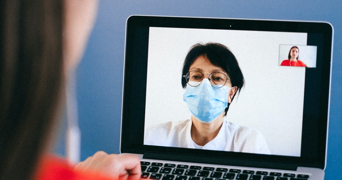 A doctor in a mask on a laptop screen speaking to a patient