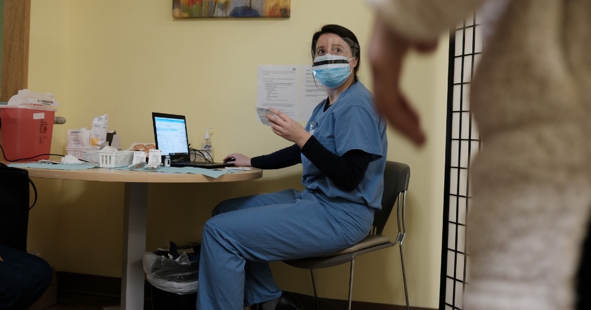 A person in scrubs and personal protective equipment sits at a computer