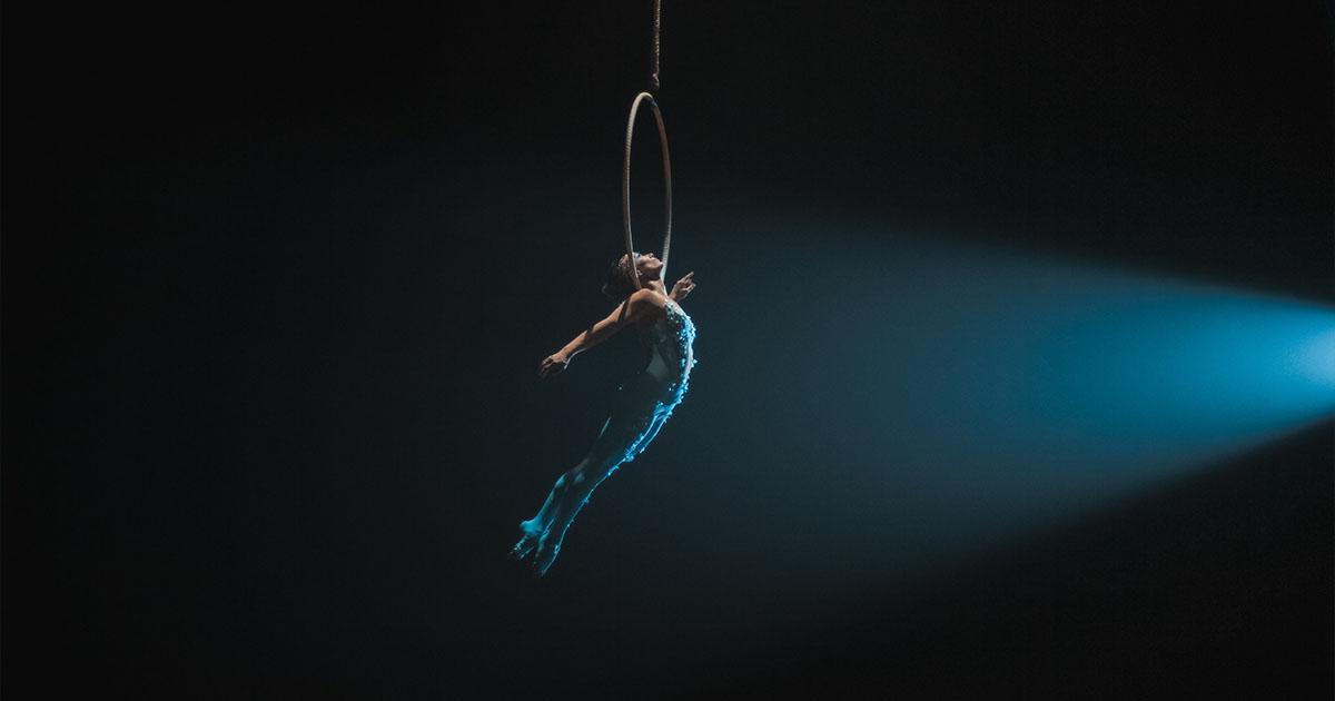 Trapeze artist in a blue costume holding onto a hoop in mid-air against a black backdrop