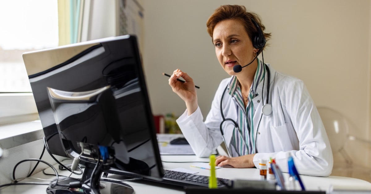 A doctor sits at a computer with a headset on