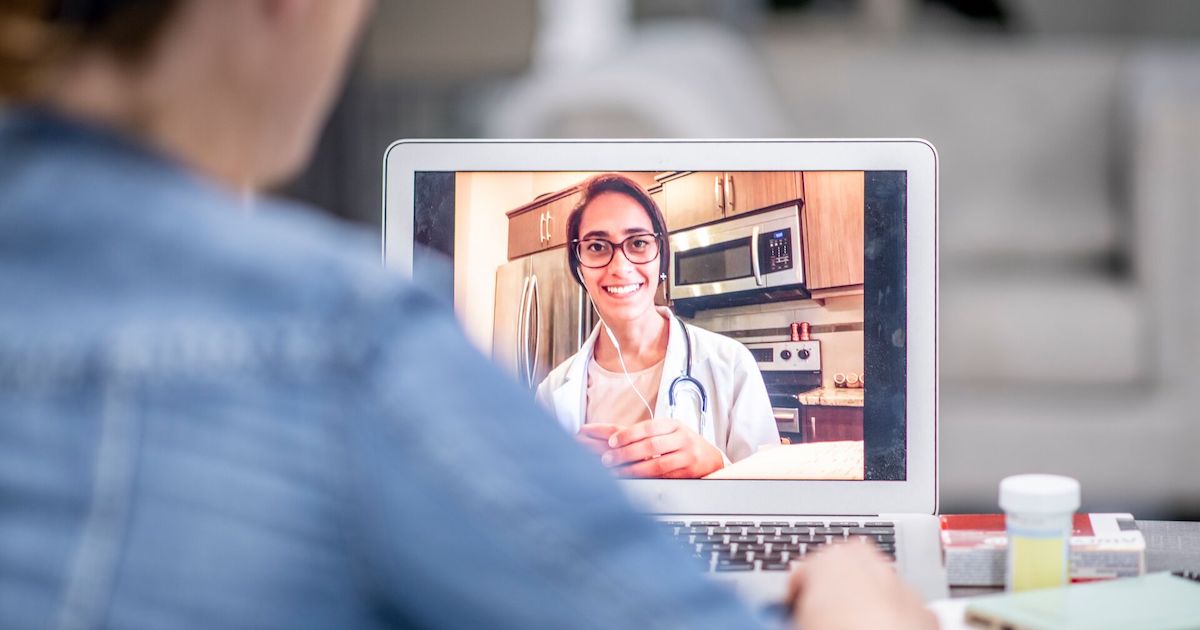 A female with a doctor's coat and glasses conducts a telehealth video call. There is a shoulder of a patient in the foreground.