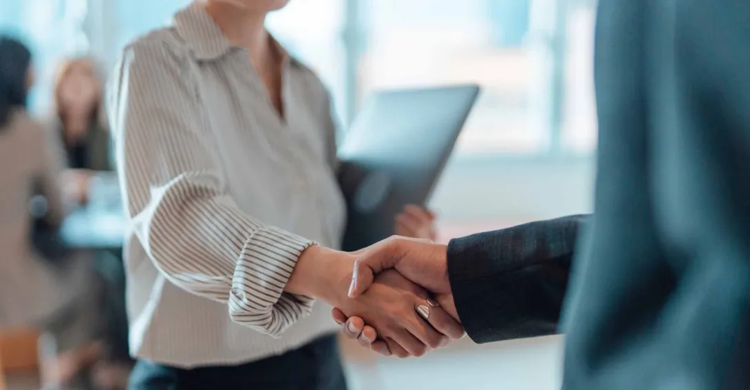 An executive in a suit shakes the hand of a person carrying a laptop