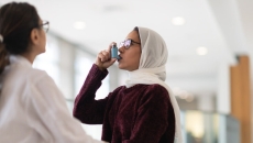 A patient uses an inhaler for asthma while a person in medical wear watches.