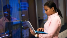 A person stands with a laptop in front of a stack of computer servers