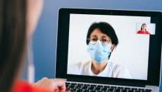 A doctor in a mask on a laptop screen speaking to a patient