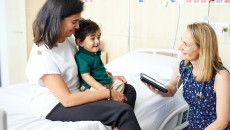 A doctor holding a tablet computer sits beside a mother and her child on a hospital bed.
