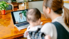 A person holding a baby and having a teleconsult with a doctor.