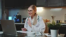 Female doctor in a kitchen waves to a virtual care patient on a laptop 