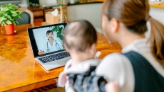 A person with a baby uses video telemedicine at a laptop