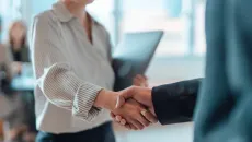 An executive in a suit shakes the hand of a person carrying a laptop