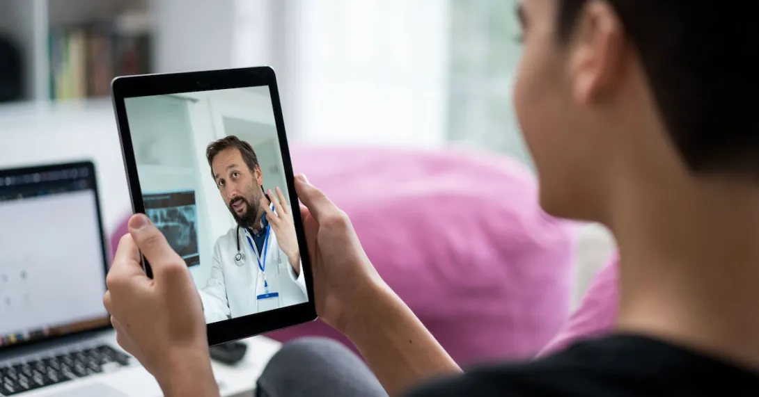 A doctor appears on a tablet having a telehealth visit with a person with short hair. A doctor appears on a tablet having a telehealth visit with a person with short hair.