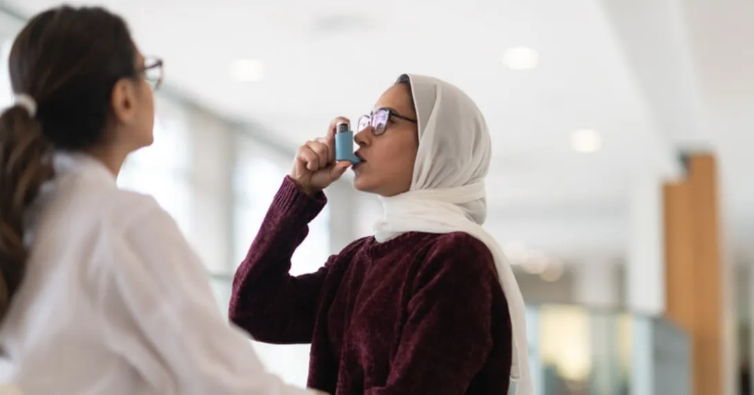 A patient uses an inhaler for asthma while a person in medical wear watches.