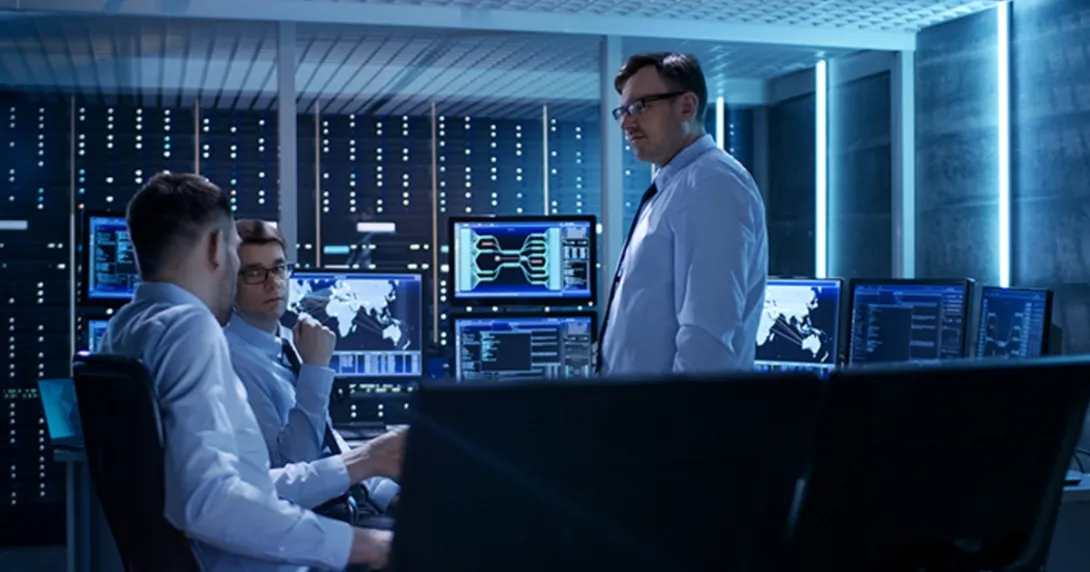 Three cybersecurity professionals talk in a network control room. Three cybersecurity professionals talk in a network control room.