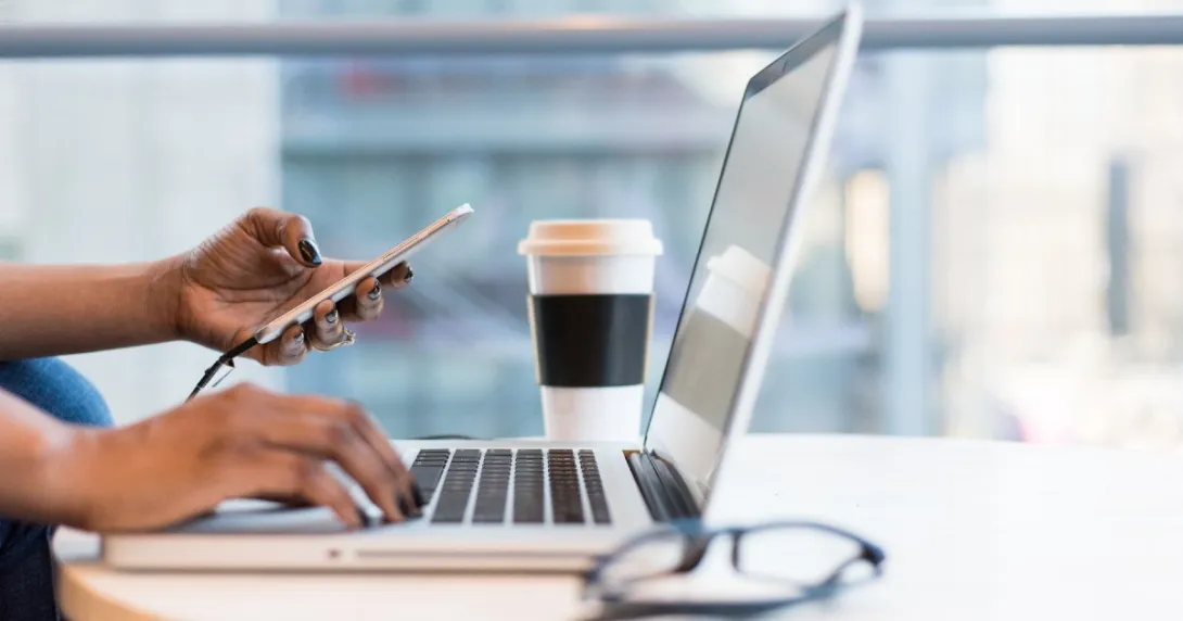 One hand rests on a laptop keyboard while the other holds a phone