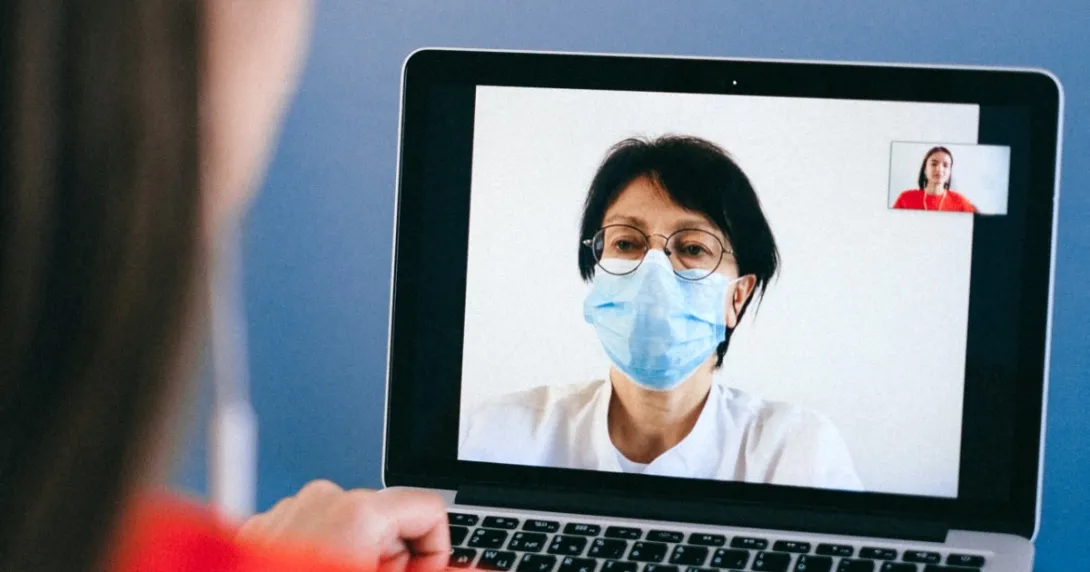 A doctor in a mask on a laptop screen speaking to a patient