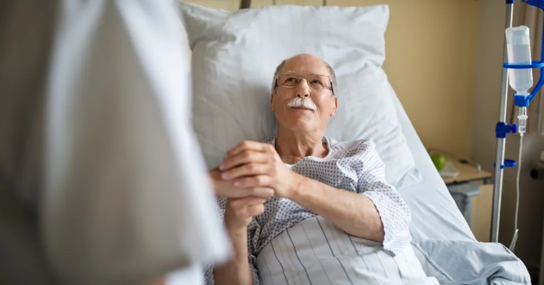 A patient in a hospital bed smiles and clasps the hand of a clinician. A patient in a hospital bed smiles and clasps the hand of a clinician.