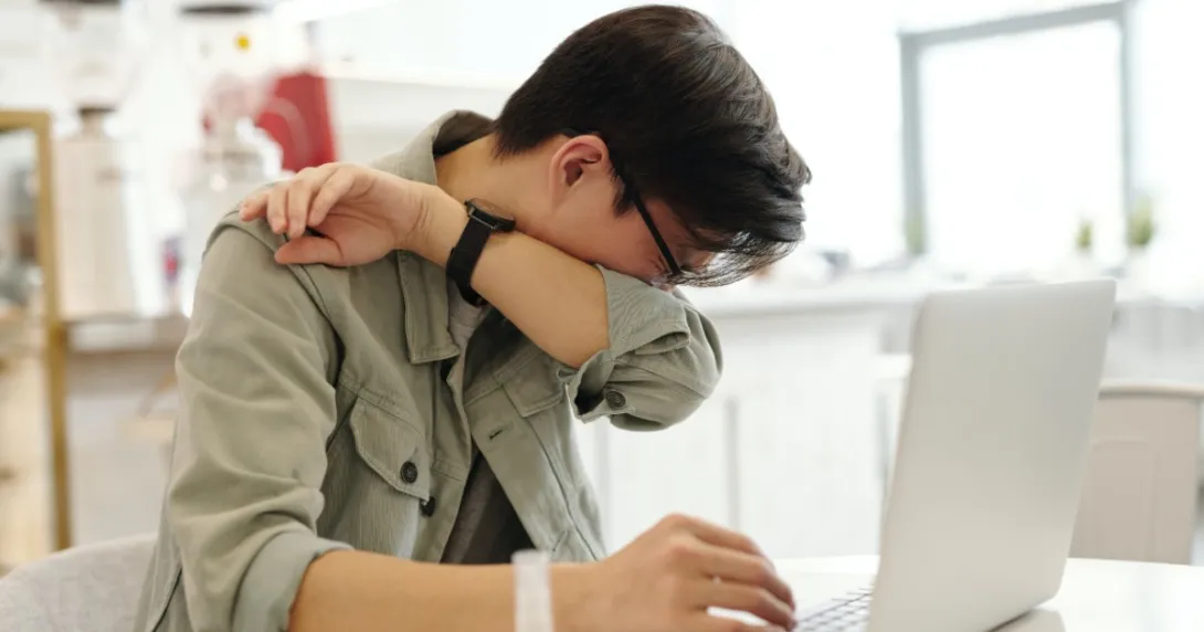 A person sneezes into their elbow in front of a computer