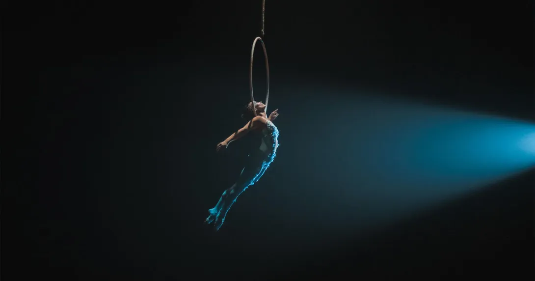 Trapeze artist in a blue costume holding onto a hoop in mid-air against a black backdrop Trapeze artist in a blue costume holding onto a hoop in mid-air against a black backdrop