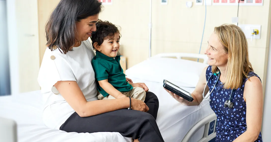 A doctor holding a tablet computer sits beside a mother and her child on a hospital bed.