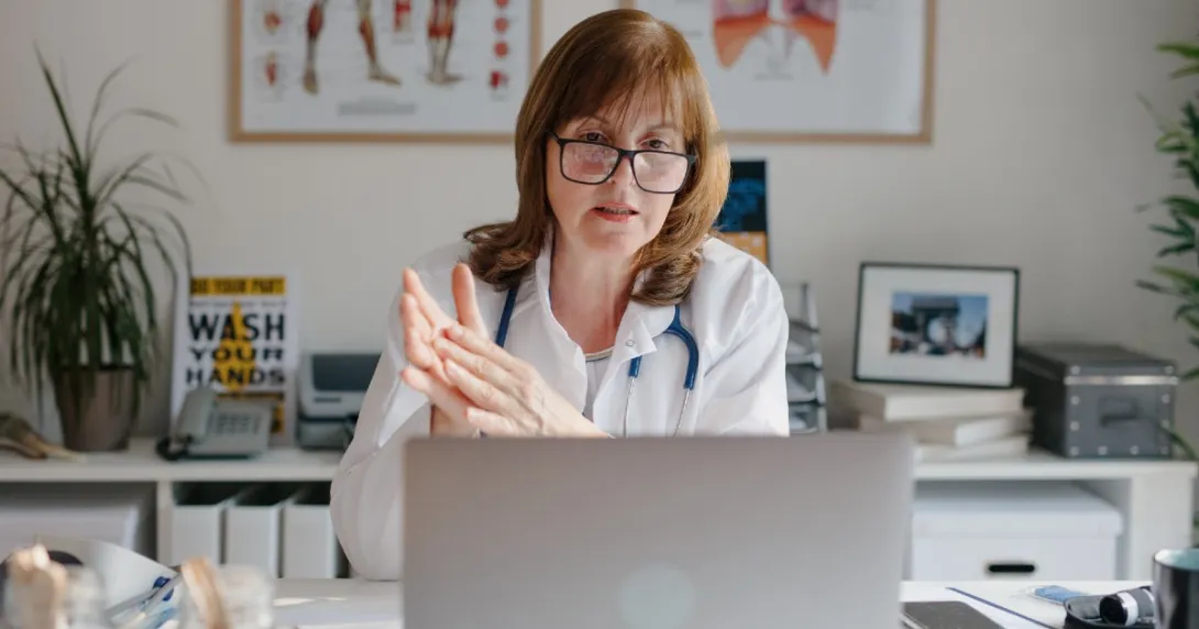 A doctor with glasses sits at a desk and speaks during a telehealth visit. A doctor with glasses sits at a desk and speaks during a telehealth visit.