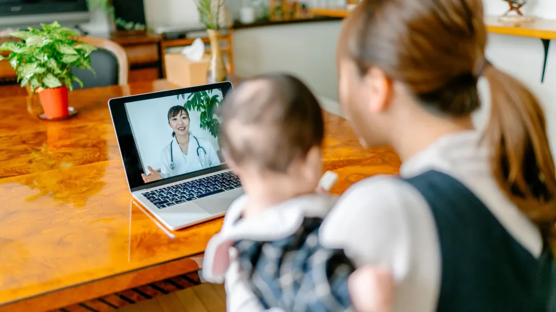 A woman holding a child while speaking with a doctor via teleconsult.