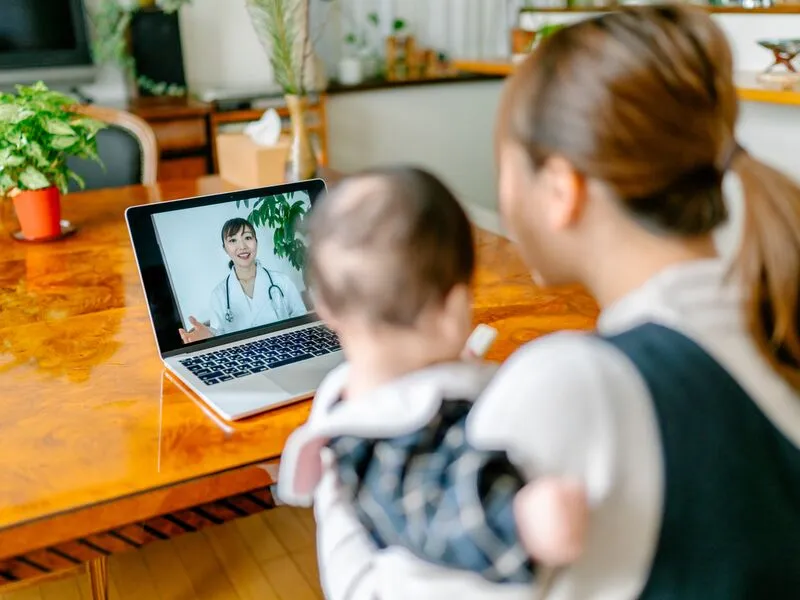 A person holding a baby and having a teleconsult with a doctor.