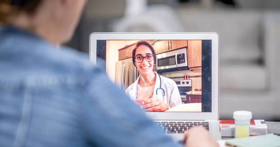 A female with a doctor's coat and glasses conducts a telehealth video call. There is a shoulder of a patient in the foreground.