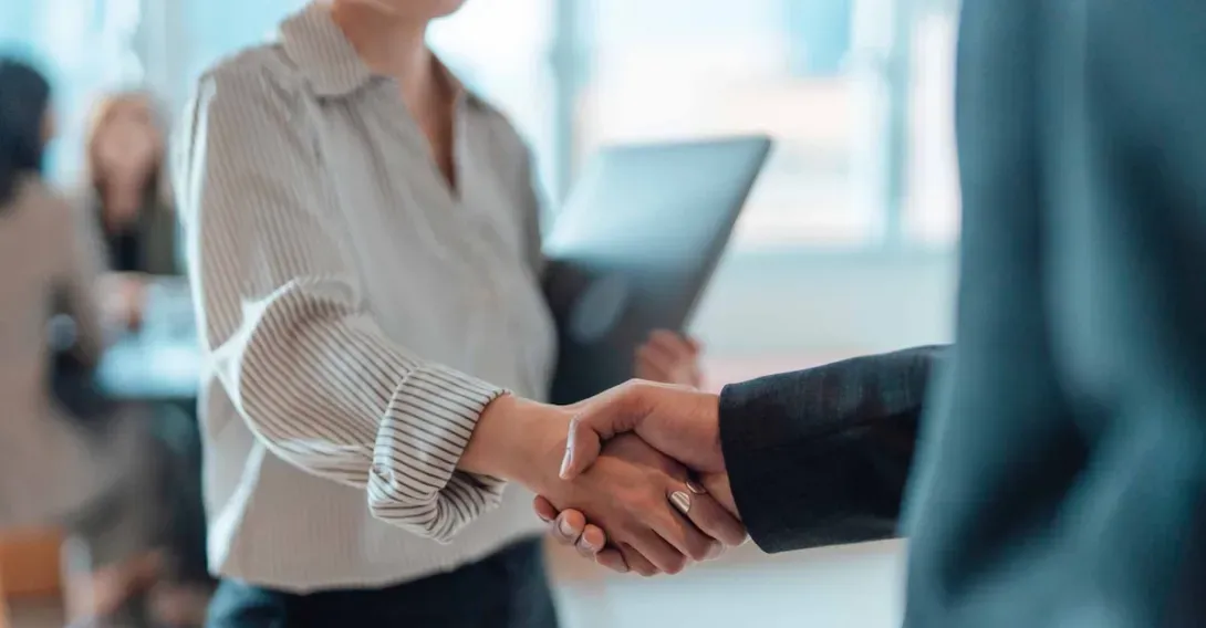 An executive in a suit shakes the hand of a person carrying a laptop