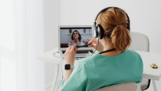 A clinician in green scrubs with red hair is conducting a virtual visit with a patient.