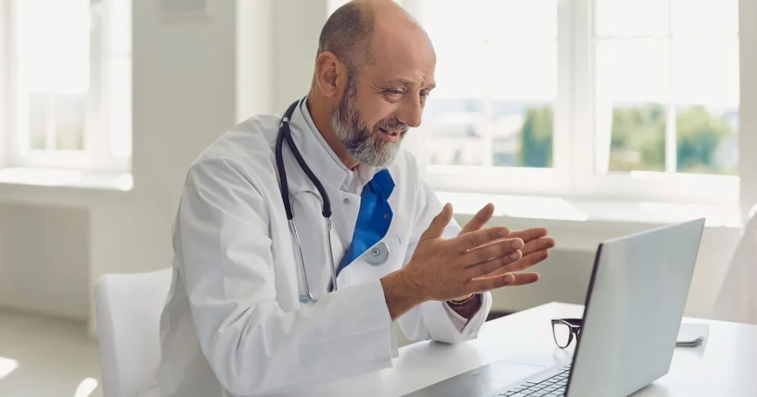 Healthcare provider with a lab coat and stethoscope sitting at a desk speaking with someone on a computer